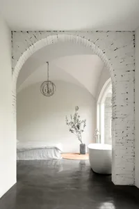 Modern minimalist bedroom with a white bed and a round tree in a vase next to a freestanding bathtub, all under a white brick