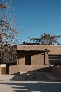 A minimalist clay exterior building with a wooden door and two stools in front of it.