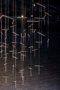 The image depicts a modern installation featuring geometric wood sculptures with attached metal rods illuminated by hanging l