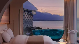 Modern hotel suite featuring a glass bathtub overlooking a lake at sunset.