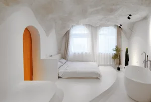 A minimalist bedroom with white walls, concrete ceiling, and floor, featuring a bed with white bedding and a bathtub near the