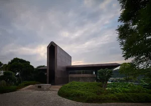 A modern house with a metal and glass exterior, surrounded by greenery and a stone pathway.