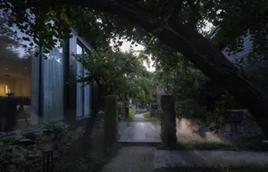 An evening view of a serene garden pathway with a stone path illuminated by mist and surrounded by lush greenery and trees.