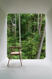 A minimalist wooden chair positioned in front of a large window, with a white wall and floor, overlooking a dense forest.
