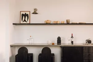 A minimalist kitchen with black stools, concrete countertop, wooden cabinets, and a wooden shelf with various ceramics and a 