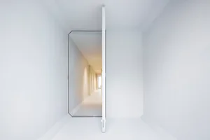 A minimalist white interior space with a glass door leading to a corridor.