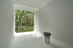 A minimalist white room with a metal stool and forest view through a large window.