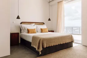 A modern bedroom with a wooden bed frame, white linens, and golden pillows, featuring a large window overlooking the sea.