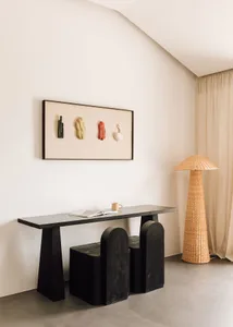 A minimalist living space featuring a sleek black console table, a wicker floor lamp, and a contemporary wall installation.