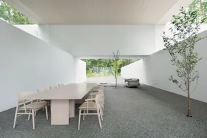 A minimalist lounge area with a long white table, chairs, and a sofa surrounded by concrete walls and floors, featuring a sma