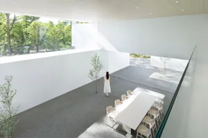 A minimalist conference room featuring a white table and chairs, surrounded by large windows overlooking greenery.