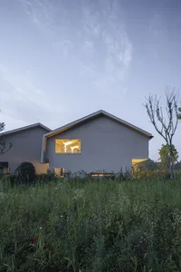 A minimalist residential concrete house with illuminated windows at dusk, surrounded by tall green grass