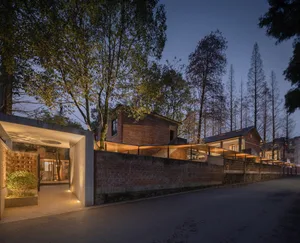 A modern residential house with brick walls and concrete walkways, illuminated by warm lighting at dusk, surrounded by trees.