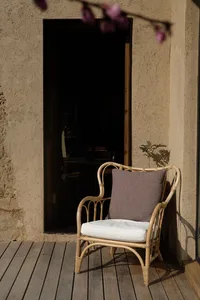 A woven bamboo chair with a white cushion and a brown pillow sitting on a wooden deck in front of a closed door.