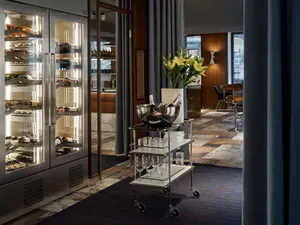 A metallic wine bar with wine bottles lined up in a glass case and a rolling cart filled with glasses and champagne bottles.