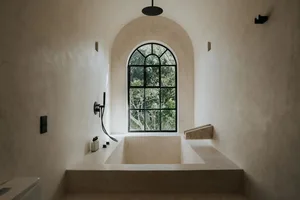 Modern bathroom featuring a concrete bathtub under a large arched window with a view of trees.