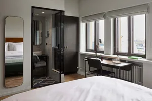 A minimalist bedroom featuring glass and wood materials, with a large bed, a desk against the window, and a bathroom visible
