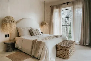 A minimalist bedroom with a textured cotton bedspread, a woven bench, and natural light from a large window overlooking green