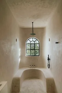A minimalist concrete bathroom with an arched window, black fixtures, and a ceiling-mounted shower head.