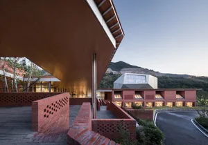 Photograph of a modern building with red brick walls and a large overhanging roof, set against a mountainous backdrop.
