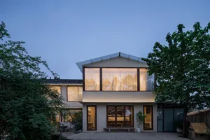 A modern residential home with large glass windows illuminated at dusk, surrounded by greenery.