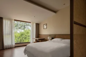 A minimalist bedroom featuring a double bed with a white textured headboard, white bedding, and large windows with sheer curt