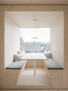 Minimalist interior with wood bench, white table, and large window overlooking rooftops and greenery.
