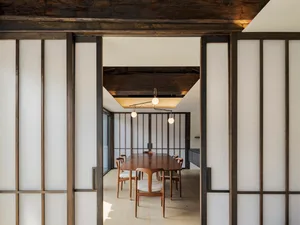 A modern dining room with wooden sliding doors and a wooden dining table surrounded by chairs. The room features a minimalist