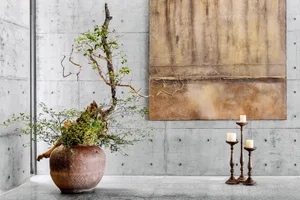 A minimalist room with a concrete floor plan, a wooden vase filled with green foliage, and two candlesticks with white candle
