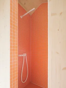 A minimalist bathroom with orange ceramic tiles on the walls and floor, featuring a white showerhead and hose.