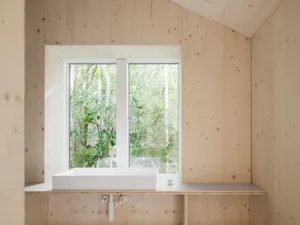 Modern minimalist bathroom with wood walls and a white sink, featuring a window with a view of the forest.