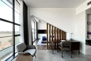 A modern minimalist bedroom with a large window, a bed with blue bedding, a wooden desk, and a comfortable seating area.