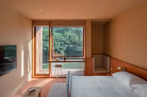 A minimalist bedroom with a large window overlooking a forest, featuring a bed with a wooden headboard and white bedding, and