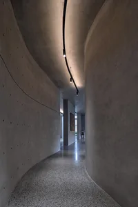 Modern concrete corridor interior with curved walls and floor, minimalistic lighting, and a view into another room.