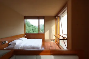 A modern minimalist bedroom featuring a wooden bed with white bedding, natural light, and a view of greenery through large wi