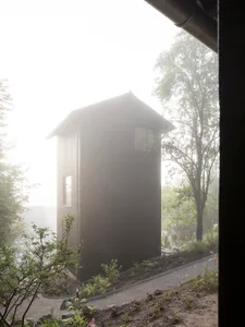 A modern minimalist house made of wood with a single window and surrounded by greenery in a foggy environment.