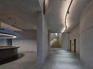 A modern office space with concrete walls and floors, featuring a curved reception desk and a staircase leading to another le
