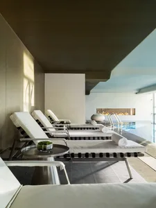 Minimalist spa lounge area with concrete walls and floor, featuring a poolside fireplace and white lounge chairs.