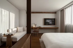 A modern bedroom with a white bed, a wooden accent wall, a white sofa, and a wooden desk with a flat-screen TV and a lamp.