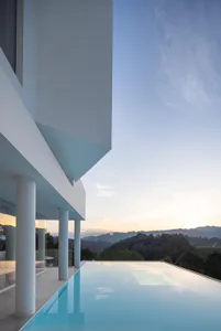 Modern, minimalist swimming pool with a glass roof and veranda overlooking mountains and sky at sunset.