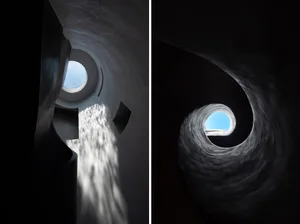 A circular, concrete staircase with a central skylight, leading upwards in a spiral pattern.
