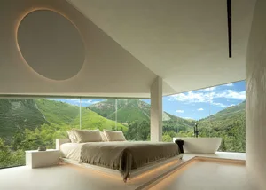 Modern bedroom featuring a concrete platform bed with a view of green hills and mountains through floor-to-ceiling windows.