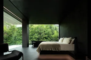 A modern bedroom featuring concrete walls with a large bed in front of a floor-to-ceiling window overlooking greenery.