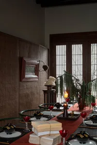 A beautifully arranged dining table with a red tablecloth, red wine glasses, black and gold bowls, and stacks of books. The r
