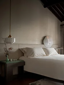 A minimalist bedroom featuring a white bed with a simple headboard, a side table with a vase and tea set, and a pendant light