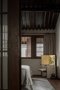 A minimalist bedroom featuring a bed with white sheets, a wooden floor, and a window with wooden shutters.