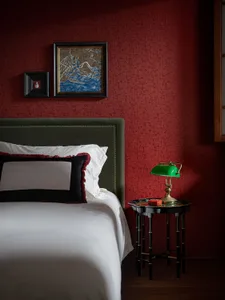 A modern bedroom with a green lamp on a bedside table, a headboard upholstered in green velvet, and a dark red textured wall.