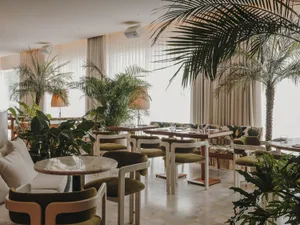 A modern restaurant interior featuring greenery, wooden tables, fabric seating, glass walls, and ceramic elements.