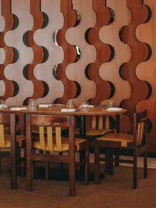 A modern restaurant interior with a wooden design featuring a round table, yellow chairs, and a decorative wave-patterned wal