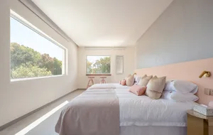 A minimalist bedroom with concrete walls, a large window, and soft pink accents on the bed and headboard.